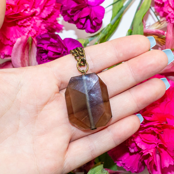 Hand holding a smoky quartz crystal pendant with pink flowers in the background