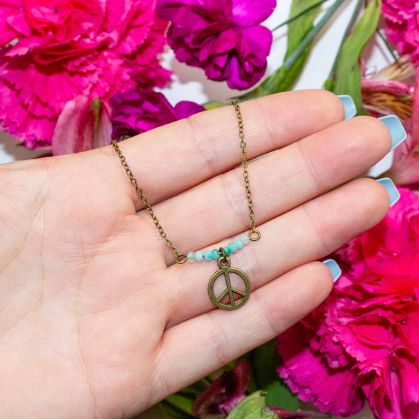 Hand holding a necklace with a peace sign pendant against a background of pink flowers