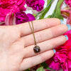 Hand holding a necklace with a smoky quartz pendant against a background of pink flowers