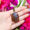 Hand holding a smoky quartz crystal pendant with pink flowers in the background