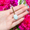 Hand wearing a necklace with a light blue amazonite pendant against a background of pink flowers