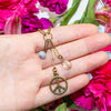 Hand holding a brass peace sign charm with pink flowers in the background